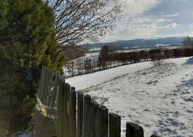 A snowy countryside view featuring a wooden fence in the foreground, with bare trees and distant hills under a clear sky.