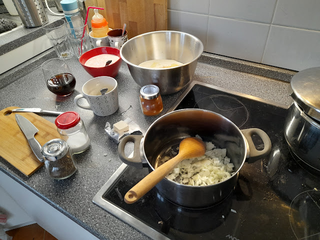 A kitchen countertop with various ingredients and utensils for making Drožďová Polievka, including chopped onions in a pot, measuring cups, containers of spices, and bowls.