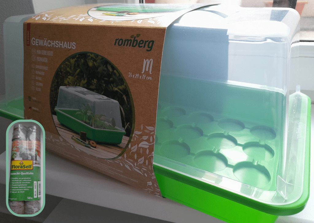 A mini greenhouse kit with a clear lid and seed starting pellets, designed for indoor gardening. The greenhouse is placed on a bright windowsill.
