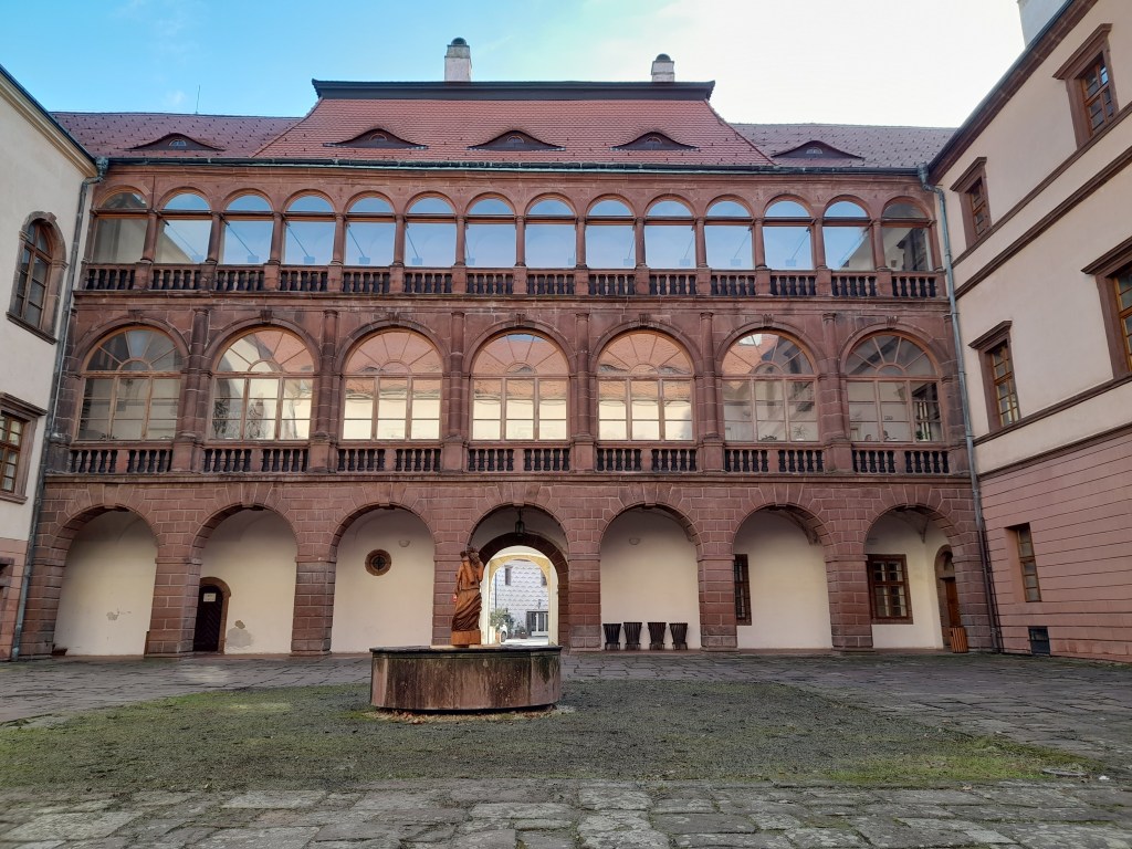 A view of the courtyard of a historical building in Kostelec nad Černými lesy, featuring arched windows and a stone fountain.