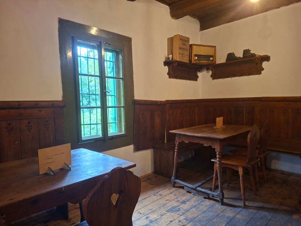 Interior of a rustic mountain hut with wooden tables and benches, a window showing greenery outside, and an old radio on a shelf.