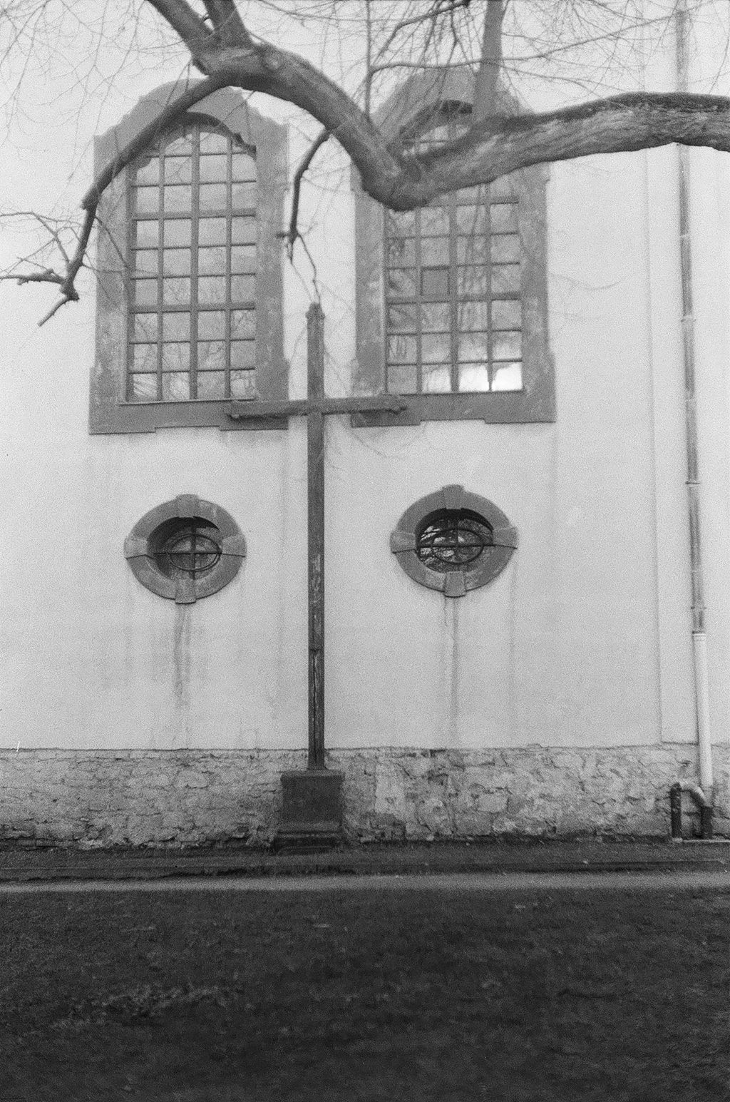 A black-and-white photograph of the side of a church featuring a wooden cross beside its wall and two large windows above, with branches from a tree extending across the top of the image.