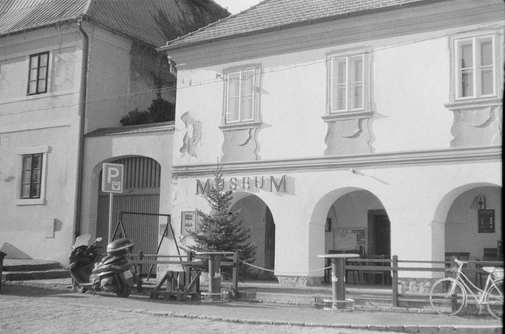 Black-and-white photograph of a museum building in Kostelec nad Černými lesy, featuring a parked scooter and a pine tree in front.