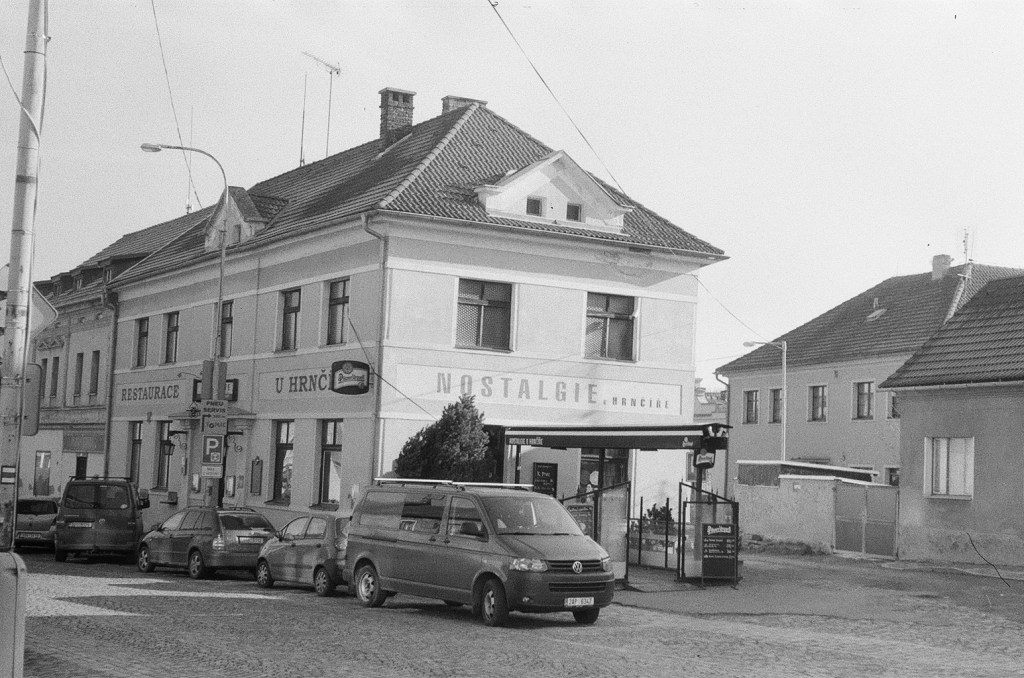 A black-and-white photograph of the Restaurant u Hrnčíře building in Kostelec nad Černými lesy, featuring the name 'Nostalgie u Hrnčíře' prominently on the facade, with cars parked in front.
