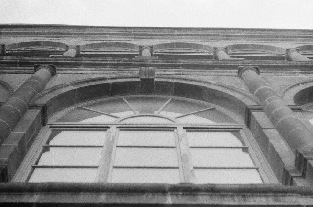 Low-angle view of a historic building's facade, featuring large arched windows and stone detailing, captured in black and white.