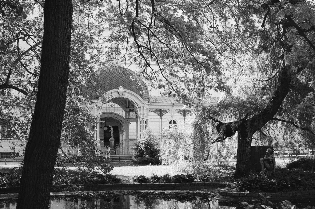 Film photograph of a female statue standing in a pond (Dvořákovy sady) with the Hygieia statue visible in the background (Sadová kolonáda); winter light, reflections on water.