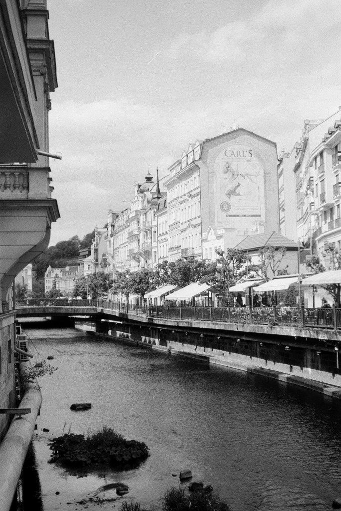 Film photograph looking down the curve of the Teplá river in Karlovy Vary, with riverside buildings and reflections.