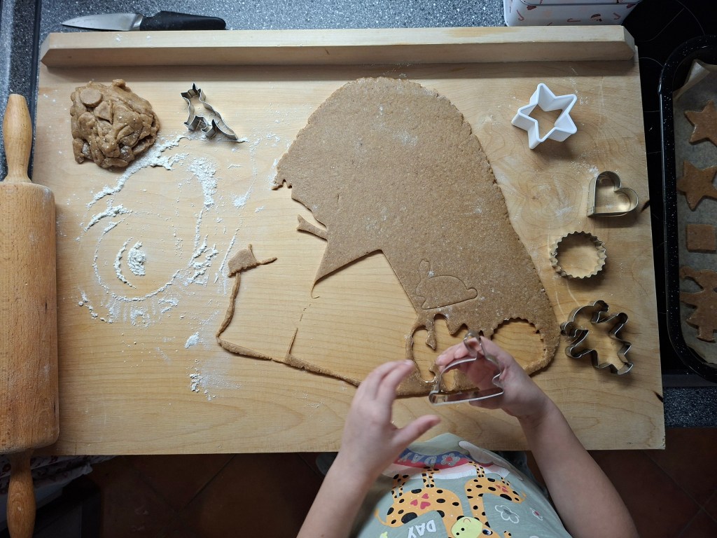 A child's hands using cookie cutters to shape gingerbread dough on a wooden surface, with flour and baking tools around.