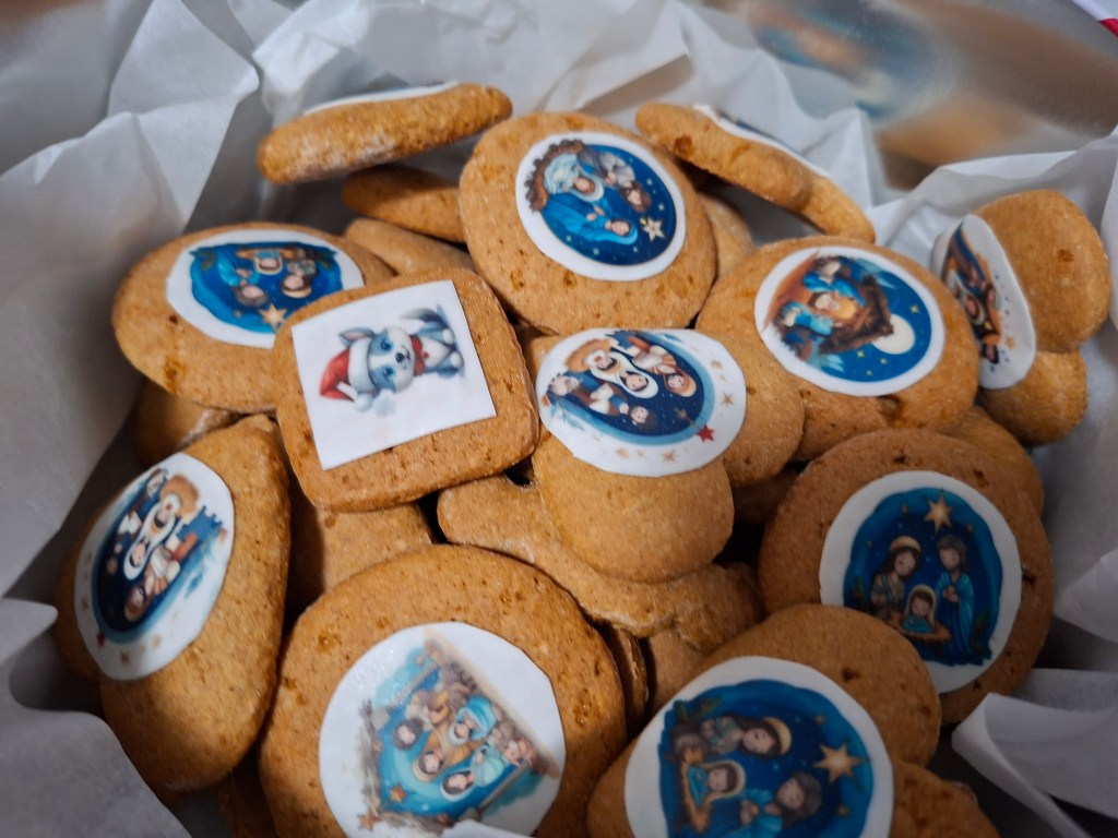 A close-up view of Christmas cookies, decorated with edible images featuring festive designs and characters, arranged in a silver container with white parchment paper.