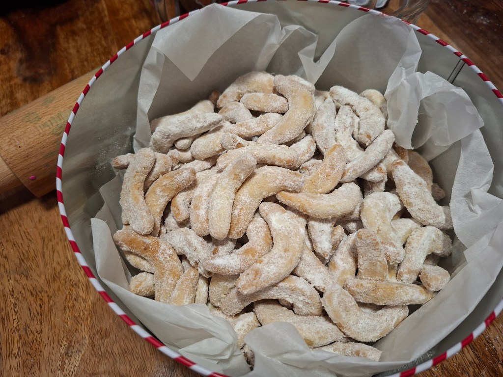 A festive tin filled with powdered sugar-coated vanilla crescents, a traditional Central European holiday cookie, resting on parchment paper.
