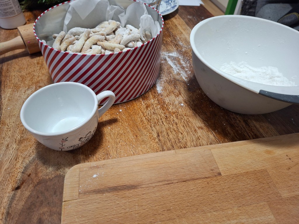 A festive kitchen scene preparing traditional vanilla crescents for Christmas, featuring a striped tin filled with cookies, a white cup on the wooden table, and a bowl of powdered sugar.