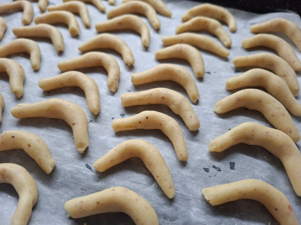 Rows of unbaked vanilla crescent cookie dough shaped into curved, small crescent forms on a parchment-lined baking tray.